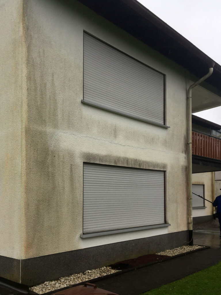 Hausfassade mit dunklen und braunen Flecken sowie Feuchtigkeitsrändern unter Fensterbänken.